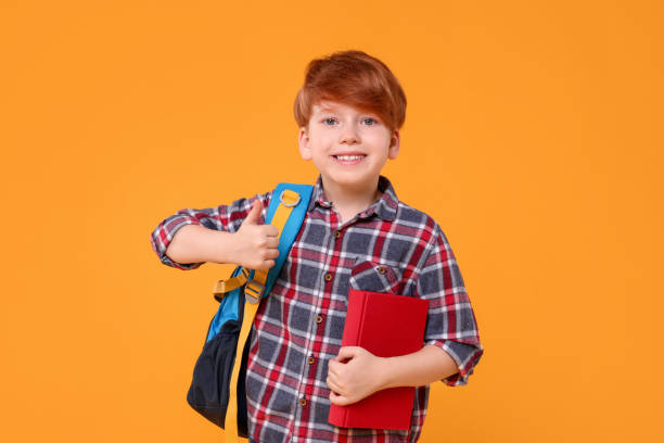 istock Happy schoolboy with backpack and book showing thumb up on orange background 1948254199