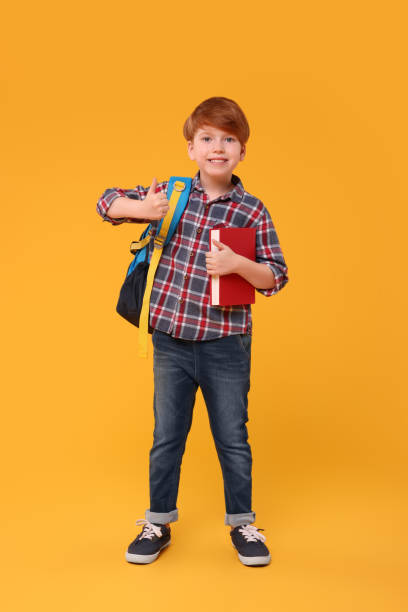 istock Smiling schoolboy with backpack and book showing thumb up on orange background 1948257921