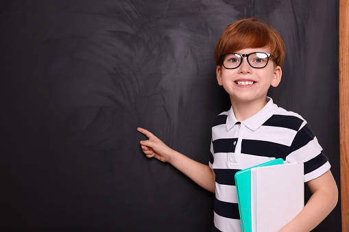 Smiling schoolboy in glasses with books pointing at something on blackboard. Space for text