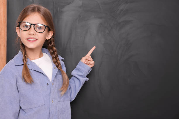 istock Smiling schoolgirl in glasses pointing at something on blackboard. Space for text 1948271183