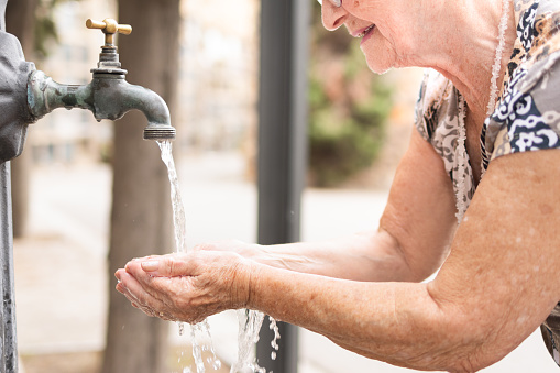 thirsty elderly woman drinking water from a fountain on the city street in a summer heat wave.