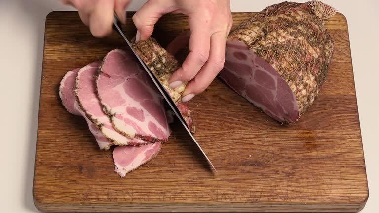 Chef slices pork ham into thin slices on a wooden board. Tasty and appetizing meat in close-up. Top view