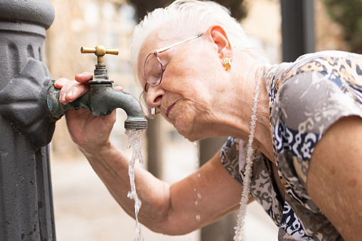 thirsty elderly woman drinking water in a summer heat wave at a public fountain on a city street. hydration and extreme temperature due to climate change.