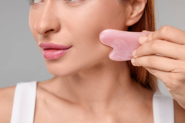 istock Young woman massaging her face with rose quartz gua sha tool on grey background, closeup 1948320137