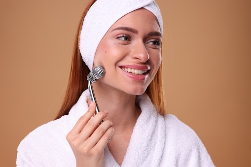 Young woman massaging her face with metal roller on pale orange background