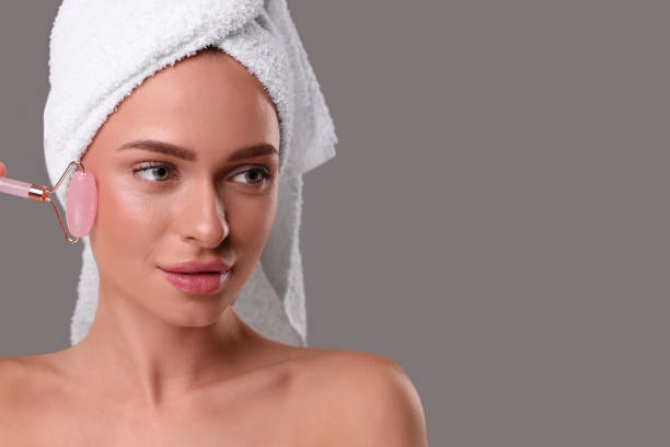 istock Young woman massaging her face with rose quartz roller on grey background, space for text 1948334337