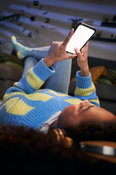 Vertical unrecognizable teenage girl lying on room carpet using mobile, blank empty screen. Black gen z woman looking cell phone with headphones at night home. Mock up concept and people domestic life