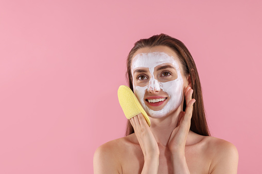 Happy young woman with face mask and sponge on pink background. Space for text