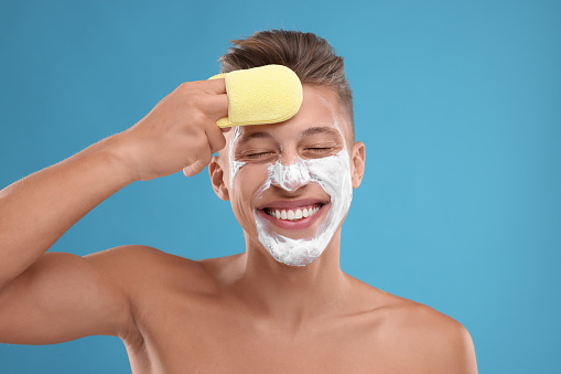 Happy young man washing off face mask with sponge on light blue background