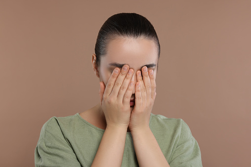 Resentful woman covering face with hands on brown background