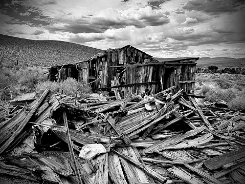 This photograph depicts a weathered and collapsing wooden structure amidst a field of debris, set against a backdrop of a hilly landscape and a cloudy sky.