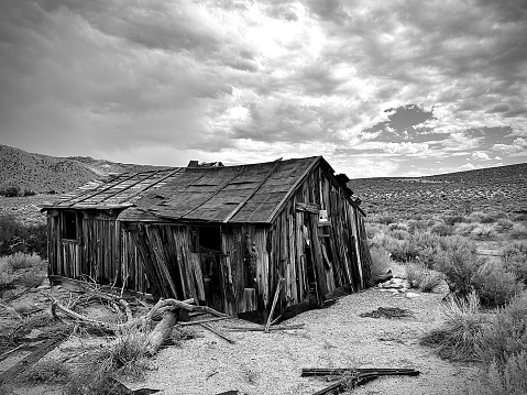 This photograph depicts a weathered and abandoned wooden shack set against a dramatic cloudy sky in a sparse desert landscape.