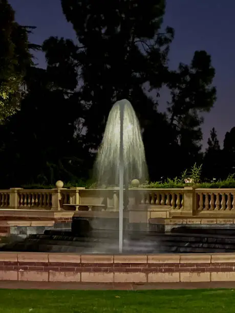 This photograph depicts a serene nighttime scene on the campus of UCLA of a fountain with a central water spout, surrounded by a balustrade and set against a backdrop of dark trees under a dusky sky.