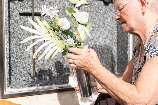 side view of sad widowed elderly woman placing flowers on family grave in cemetery