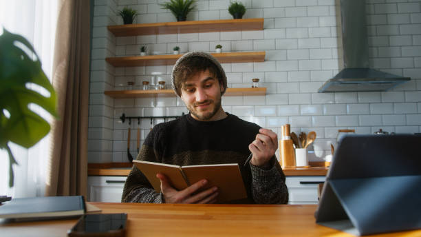 istock Smiling student with beanie sitting in modern kitchen at home looking at the notes in notebook he is holding in his hand, reading, thinking and changing the pages 1948808400