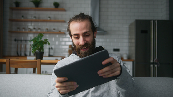 Young bearded man with hair bun sitting on sofa at home with kitchen background, playing excited concentrated engrossed in interesting mobil game with tablet computer