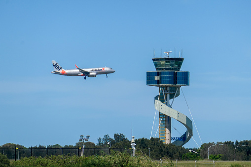 A Jetstar Airbus A320-232 plane, registration VH-YXS, on her landing approach to the third runway of Sydney Kingsford-Smith Airport as flight JQ413 from the Gold Coast.  She is about to pass the air traffic control tower.  This image was taken from Kyeemagh, Botany Bay, on a hot and sunny afternoon on 19 January 2024.