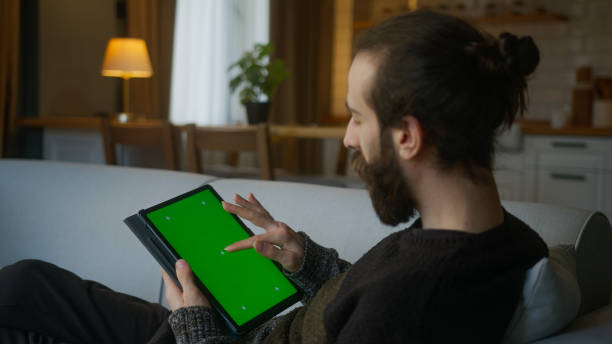 istock Bearded man scrolling and tapping center on tablet with green screen mock up display. Male lyin on sofa, relaxing at home. Close-up over the shoulder shot 1948841168
