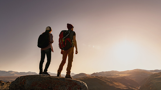 Two young active silhouettes of hikers with backpacks are standing on big rock and looks at sunset in mountains
