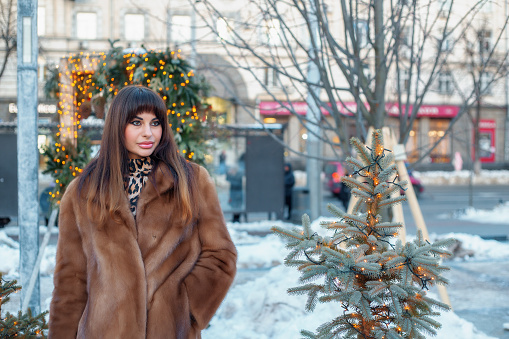 Brunette woman in a fur coat standing on a city street with a hand in her pocket looking to the side