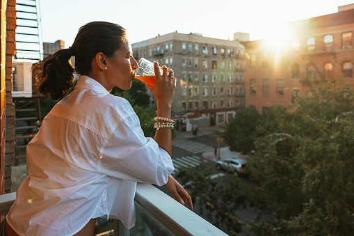 Latin woman from her house in New York look out the balcony while enjoying a refreshing drink at sunset.