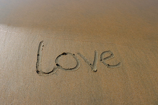 Stock photo showing writing drawn on sunny beach with word 'love' written in sand with stick in soft golden sand on seaside coastline.