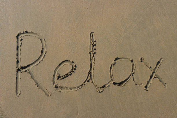 Stock photo showing writing drawn on sunny beach with word 'relax' written in sand with stick in soft golden sand on seaside coastline.