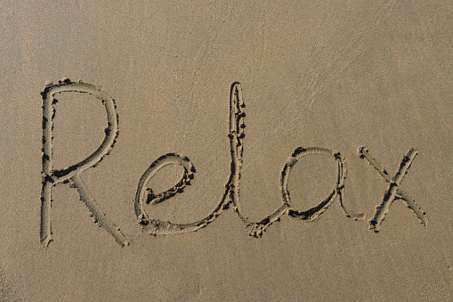 Stock photo showing writing drawn on sunny beach with word 'relax' written in sand with stick in soft golden sand on seaside coastline.