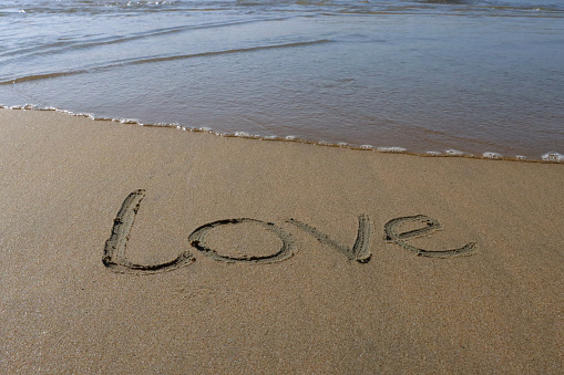 Stock photo showing writing drawn on sunny beach with word 'love' written in sand with stick in soft golden sand on seaside coastline.