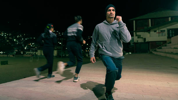 A mid-adult man does squat exercises for his fitness routine outdoors at night on a community ground while other people jog in the background.