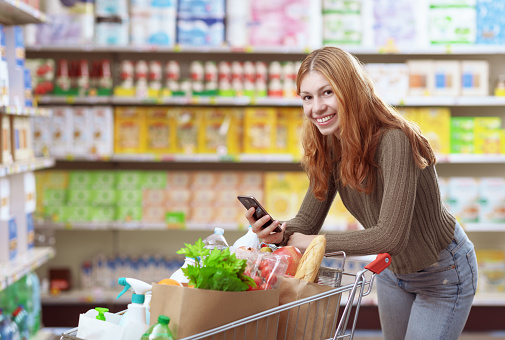 Portrait of a young woman doing grocery shopping at the supermarket, she is smiling and holding her smartphone
