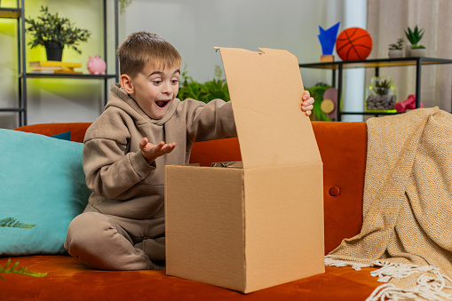 Happy excited shocked child boy unpacking delivery parcel. Smiling satisfied teenager kid shopper, online shop customer opening cardboard box receiving purchase gift by fast postal shipping at home