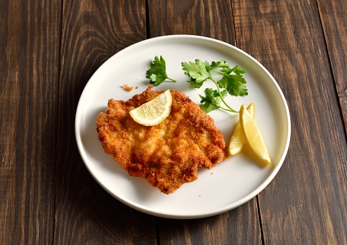 Pork schnitzel with lemon and leaves of parsley on white plate over wooden background. Close up view