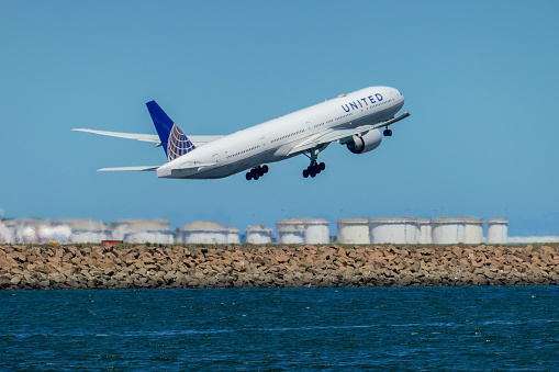 A United Airlines Boeing B777-332(ER) plane, registration N2341U, taking off to the south from Sydney Kingsford-Smith Airport and heading to San Francisco as flight UA870. In the distance are fuel storage tanks at the Port Botany Container Terminal.  This image was taken from Kyeemagh, Botany Bay, on a hot and sunny afternoon on 19 January 2024.