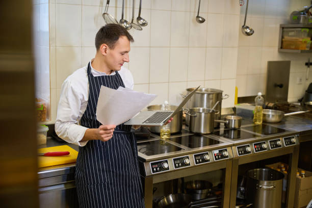 istock Kitchen employee sits with laptop and work papers at stove 1949377864