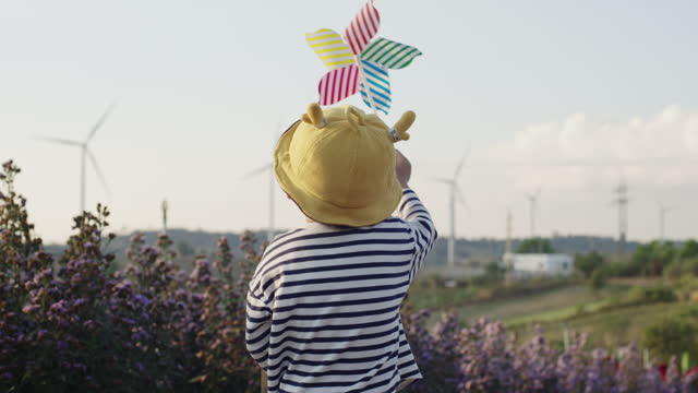 Adorable cute Asian toddler boy holding pinwheel toy wind turbine walking along beautiful outdoor flower field with wind turbine energy farm in background. Baby and kids outdoor exploration imagination learning sustainable energy lifestyle concept.