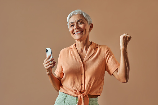 Adorable  cheerful sincere mature beautiful gray haired woman in pastel orange shirt and green pants holding phone and making win win gesture being happy and laughing. Victory, celebration, winning.