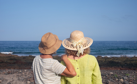 Back view of two caucasian women embracing standing at the beach face the sea looking the horizon. Love, comfort and friendship concept