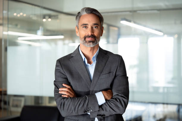 Handsome hispanic senior business man with crossed arms smiling at camera. Indian or latin confident mature good looking middle age leader male businessman on blur office background with copy space. stock photo