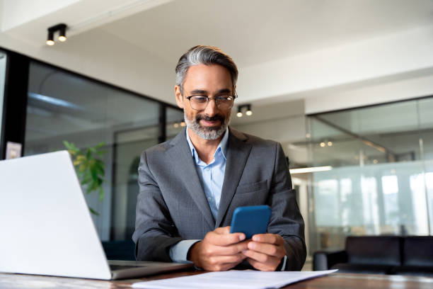 istock Middle aged Hispanic business manager ceo using cell phone mobile app, laptop. Smiling Latin or Indian mature man businessman holding smartphone sit in office working online on gadget with copy space. 1949502290