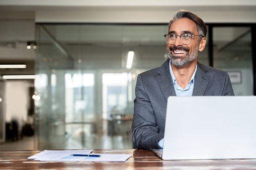 Portrait of mature Indian or Latin business man ceo trader using laptop computer, typing, working in modern office. Middle-age Hispanic smiling handsome businessman entrepreneur looking aside,dreaming