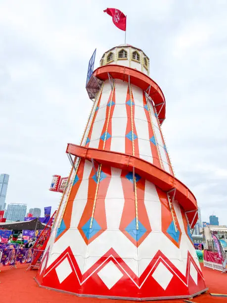 This vibrant helter-skelter, adorned with bold red and white stripes and blue accents, stands tall against a cloudy sky, inviting thrill-seekers for a spiraling descent amidst the bustling atmosphere of a lively fairground.