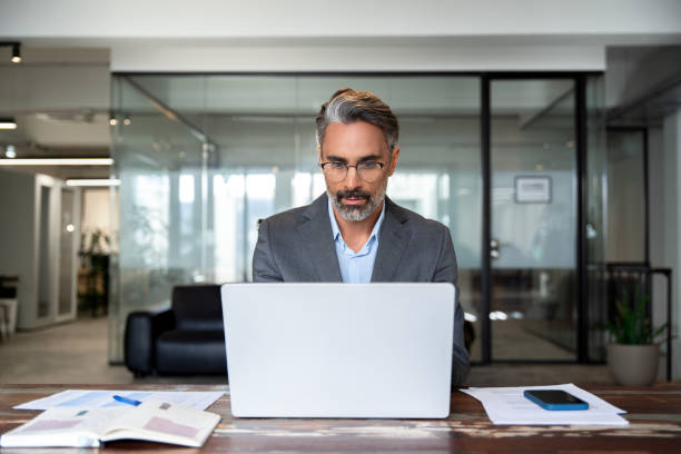 istock Mature Indian or Latin business man ceo trader using computer, typing, working in modern office, doing online data market analysis, thinking planning tech strategy looking at laptop with copy space. 1949511876