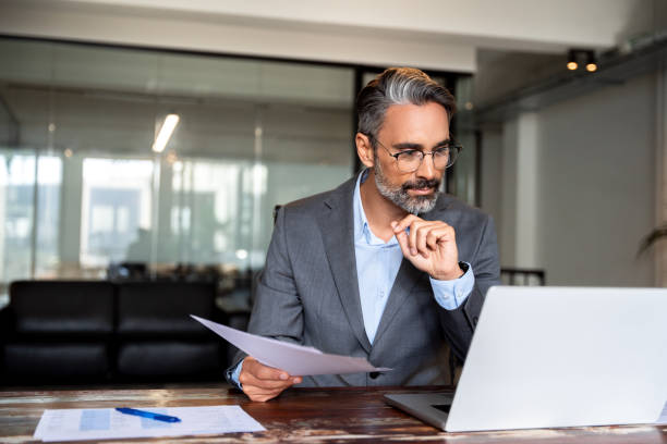 Focused Hispanic businessman with paperwork in office. Concentrated European male business man accountant analyst holding documents, work at laptop computer doing online trade market tech research. stock photo