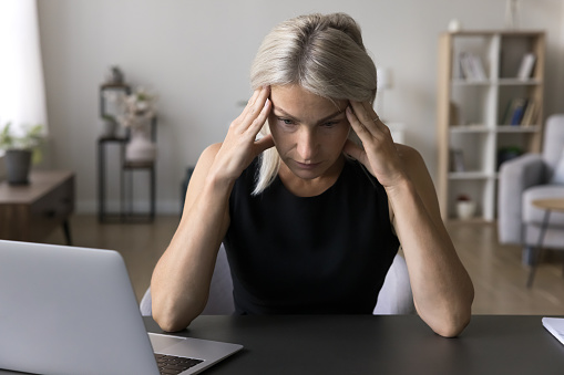 Anxious, nervous serious businesswoman sit at desk with laptop, touch forehead, search issue solution, looks overloaded due too long computer usage. Business problems, concerns, information overload