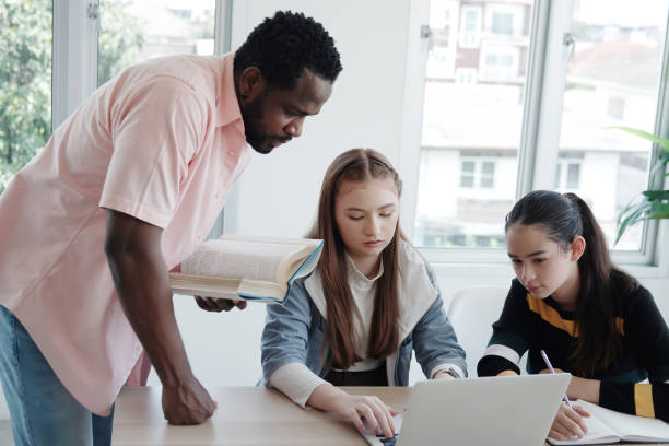istock Young African teacher or tutor is holding book and teaching for knowledge about online research with internet on laptop for Caucasian student girls in classroom. Education and Technology Concept 1949519798