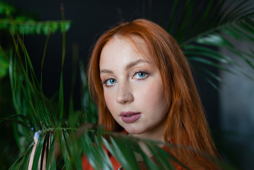 Young red hair woman florist posing at workplace with green potted plants background. Looking ar camera.