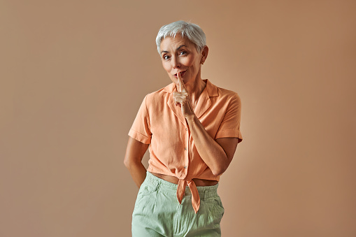 Attractive european middle aged woman dressed in peach shirt holding forefinger near lips and looking at camera asking for silence. Isolated over beige background. Copy space.Keep silence, shh.