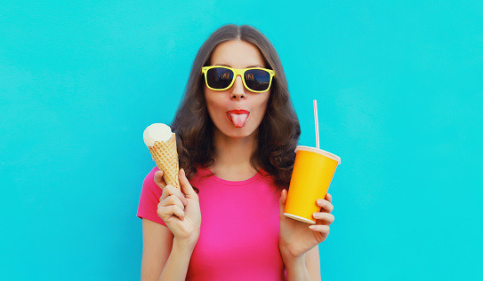 Summer portrait of happy young woman eating ice cream and holding cup of fresh juice wearing sunglasses on blue background