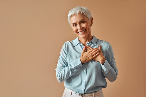 Beautiful confident woman in blue blouse holding hands over heart while standing on beige background and smiling.